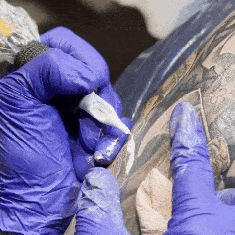 Close-up of a tattoo artist wearing purple gloves applying white ink to a tattoo on a person's arm.