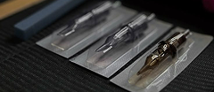 Three tattoo machine needles in individual sealed transparent plastic packages, arranged diagonally on a dark surface.
