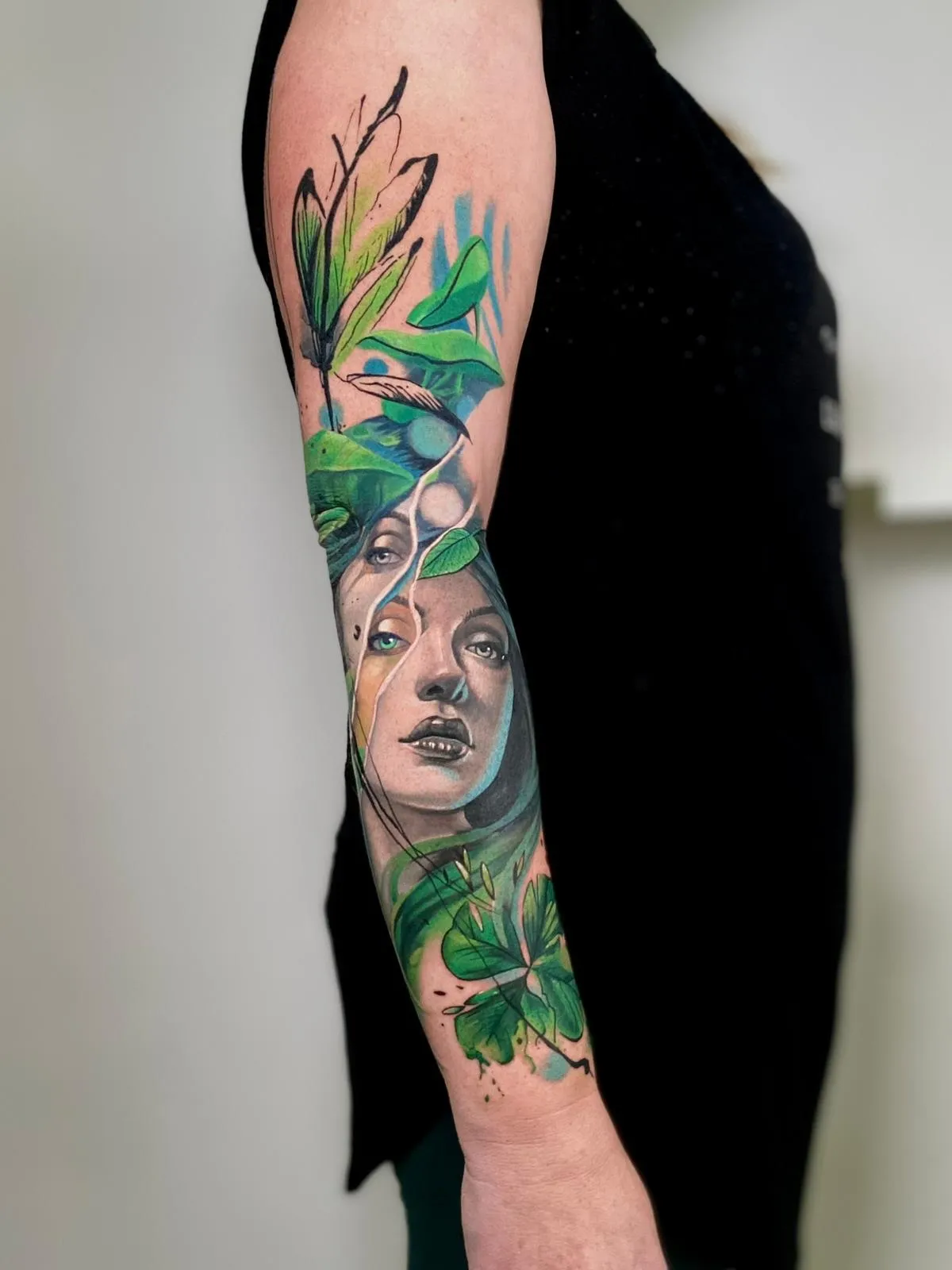 Colorful forearm tattoo featuring a realistic female face with green eyes, surrounded by green leaves, feathers, and abstract blue and black accents.