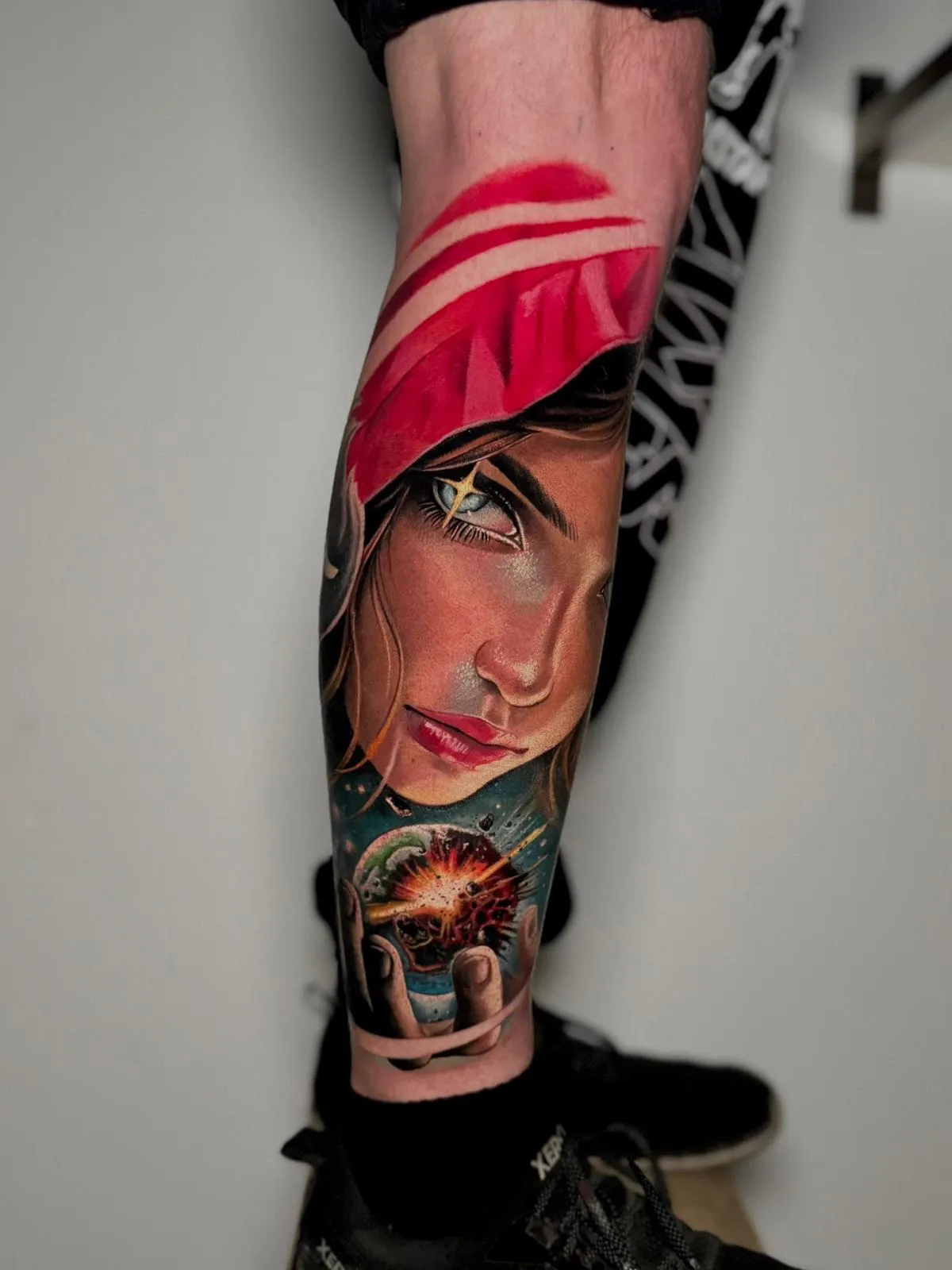 Colorful tattoo on a calf featuring a woman's face with a red hood and a glowing orb with an explosion held in her hand.