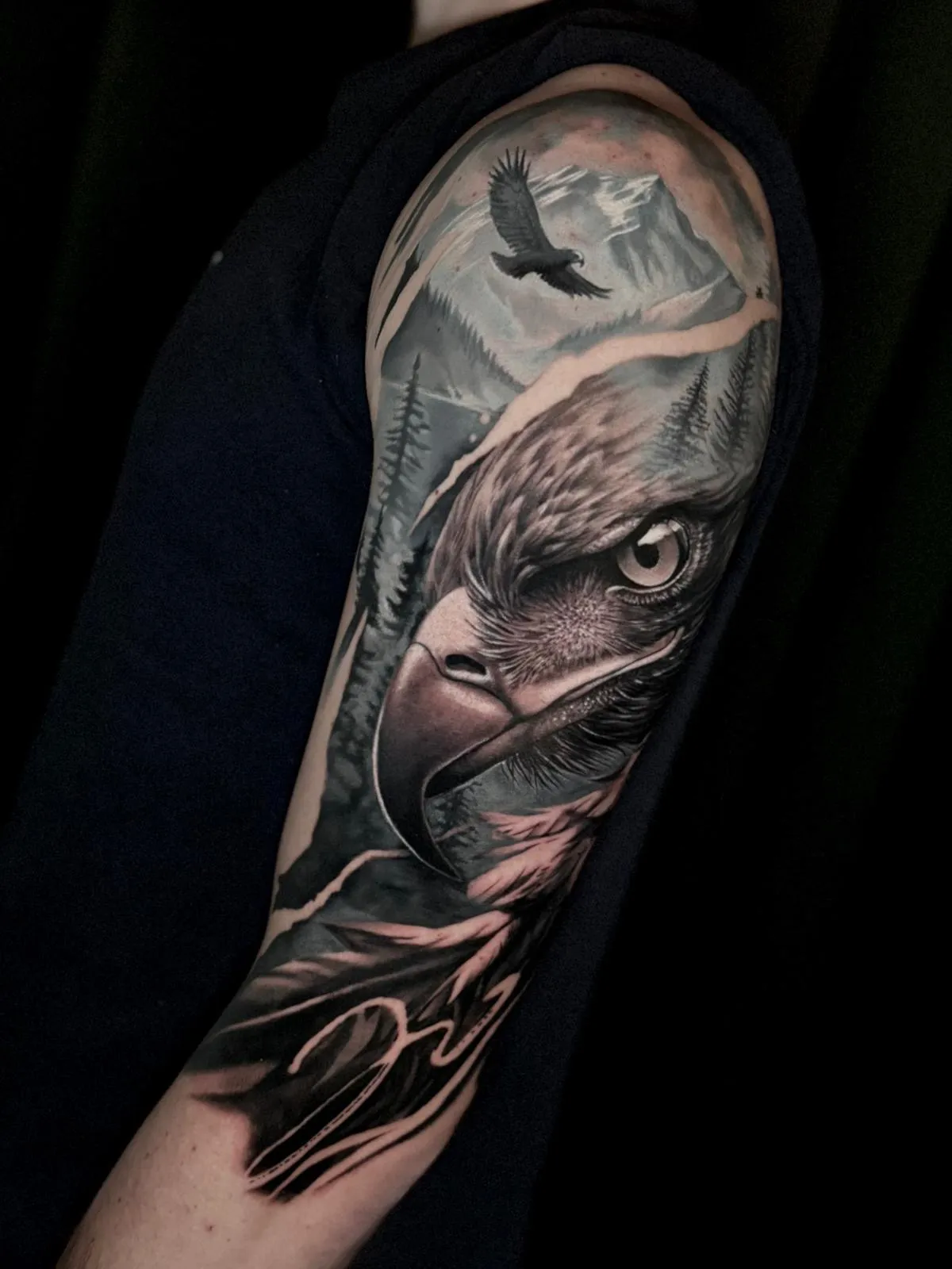 Realistic black and grey tattoo of a bald eagle head with intense eye, flying eagle, pine trees, and mountains on an arm.