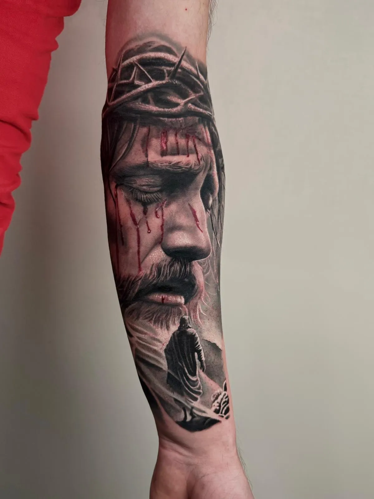 Realistic tattoo of Jesus with a crown of thorns and blood dripping down his face, with a smaller figure in a cloak walking below.