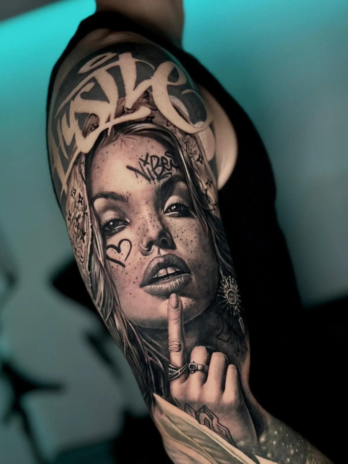 Black and gray tattoo of a woman with freckles, heart and script face tattoos, holding a finger to her lips in a shushing gesture.