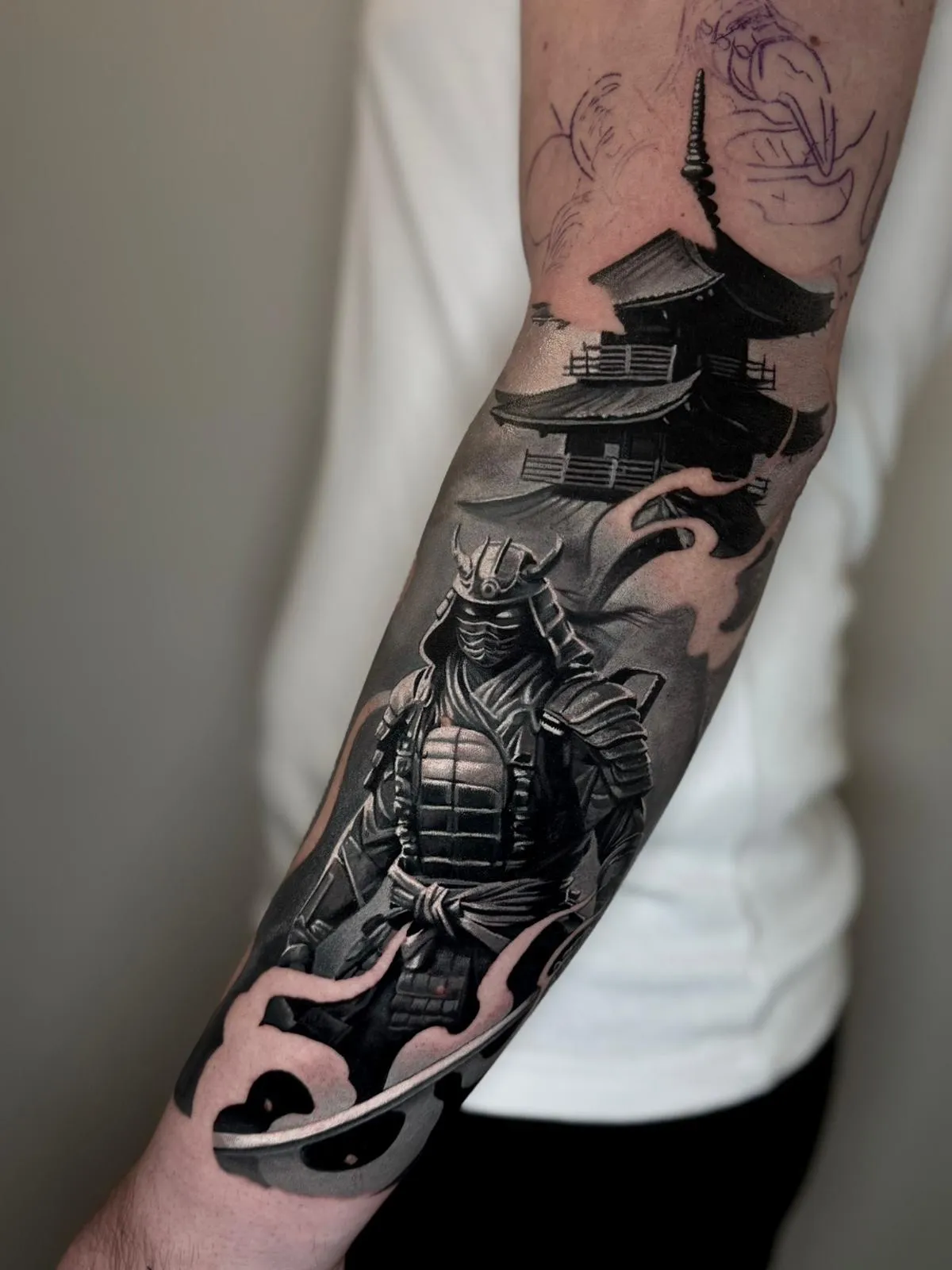 Black and grey tattoo of a samurai warrior in armor with a sword and a Japanese pagoda temple in the background on a forearm.