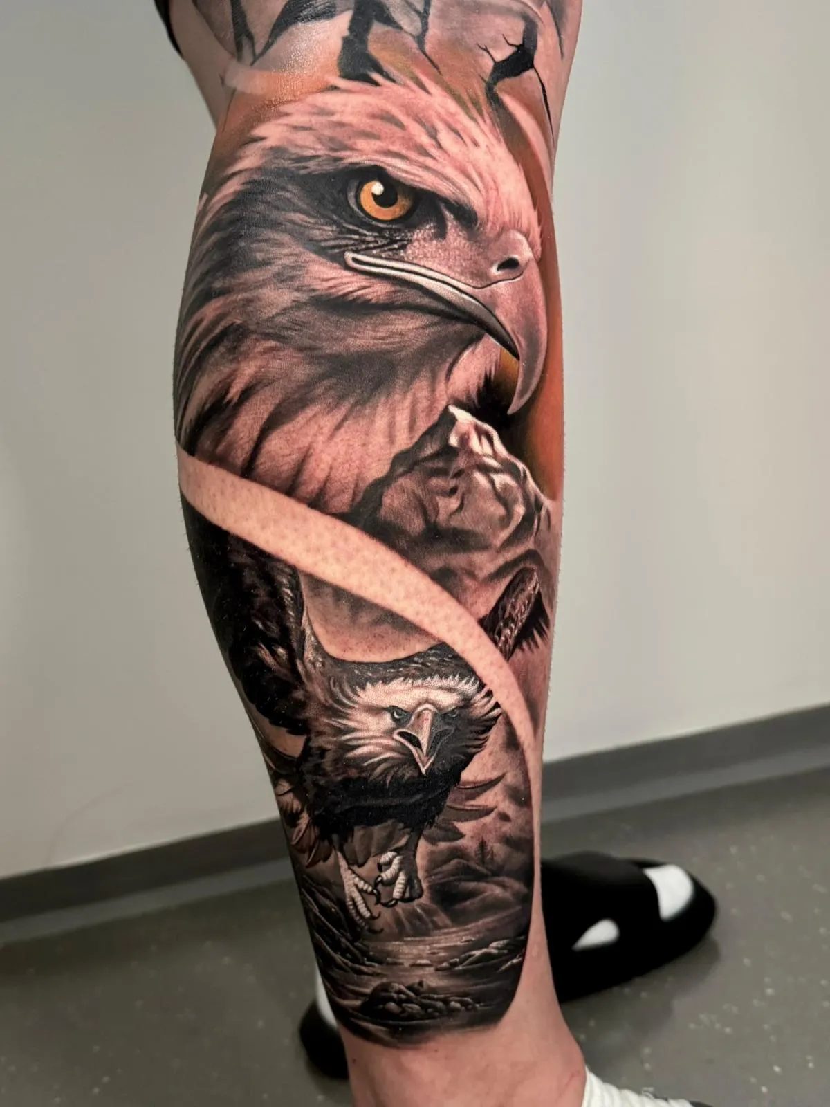 Realistic tattoo on a leg featuring a large eagle head, a roaring bear, and an eagle in flight over a mountainous landscape.
